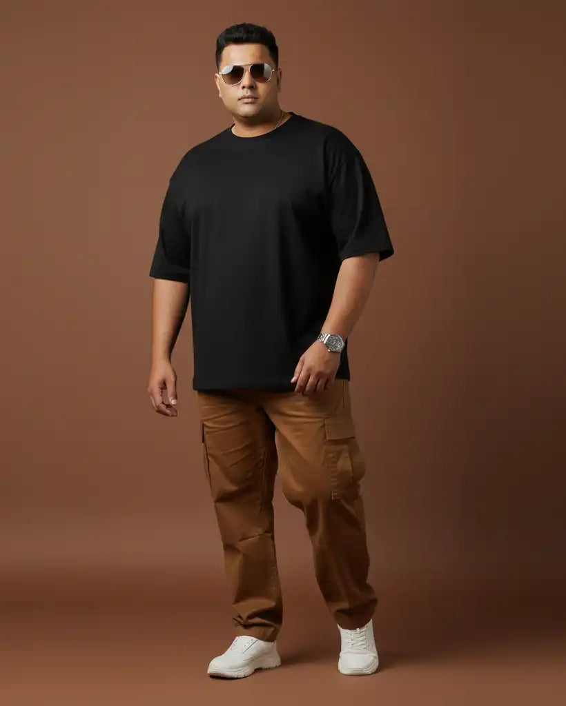 Plus size black oversized t-shirt on plus size male model in brown cargo pants and white sneakers, full front view against brown background, premium AWBlack plain tee