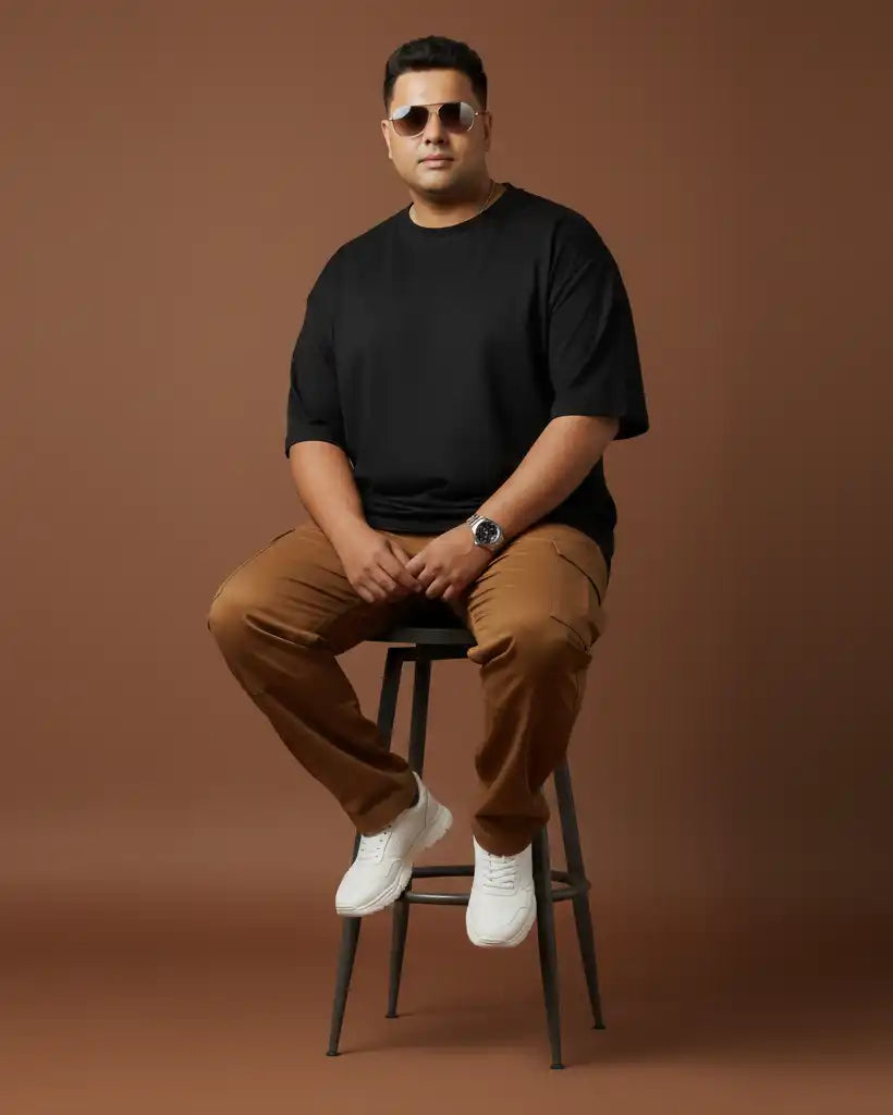 Plus size black oversized t-shirt on plus size male model sitting on stool wearing sunglasses, brown cargo pants and white sneakers, front view against brown background, premium AWBlack plain tee